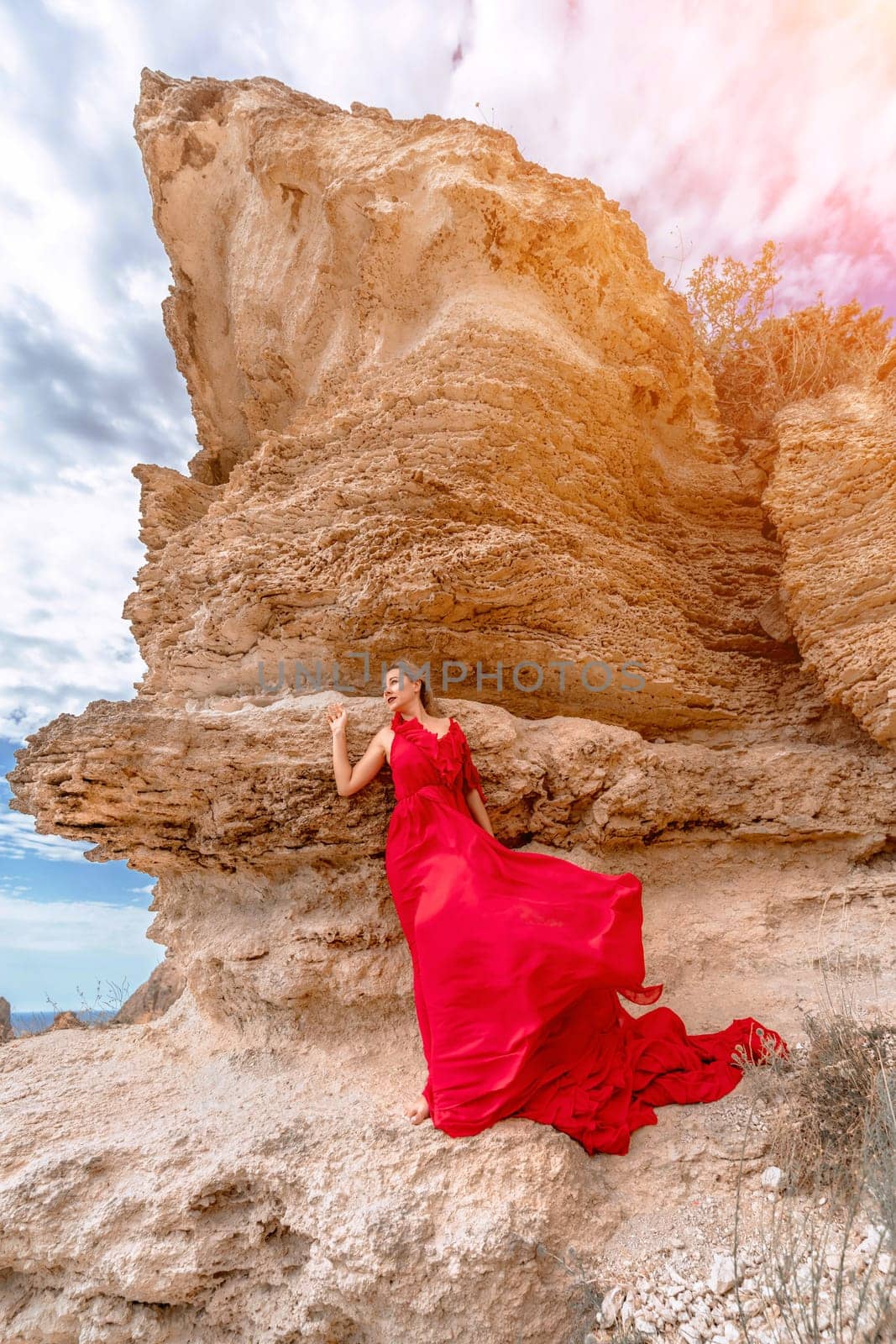 woman red silk dress stands by the ocean, with mountains in the background, as her dress sways in the breeze