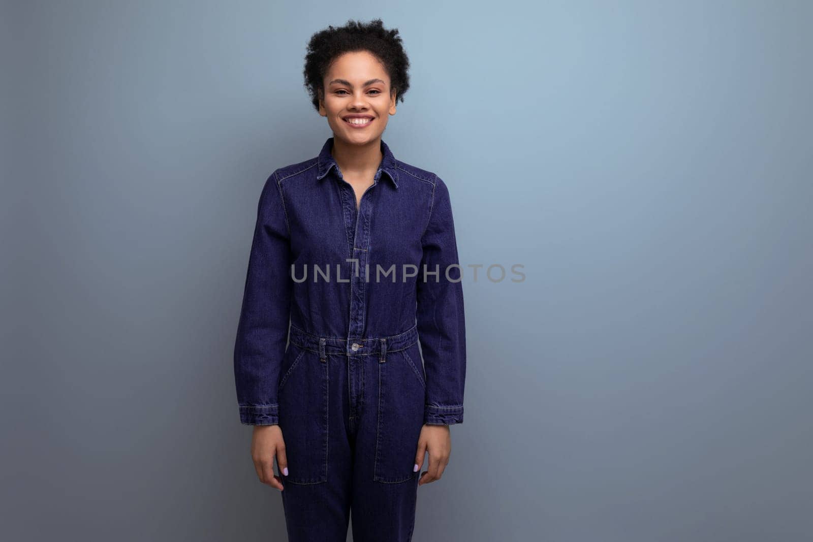 young positive brunette latin woman with afro hair gathered in a ponytail dressed in a blue stylish suit consisting of one-piece jeans and a shirt.