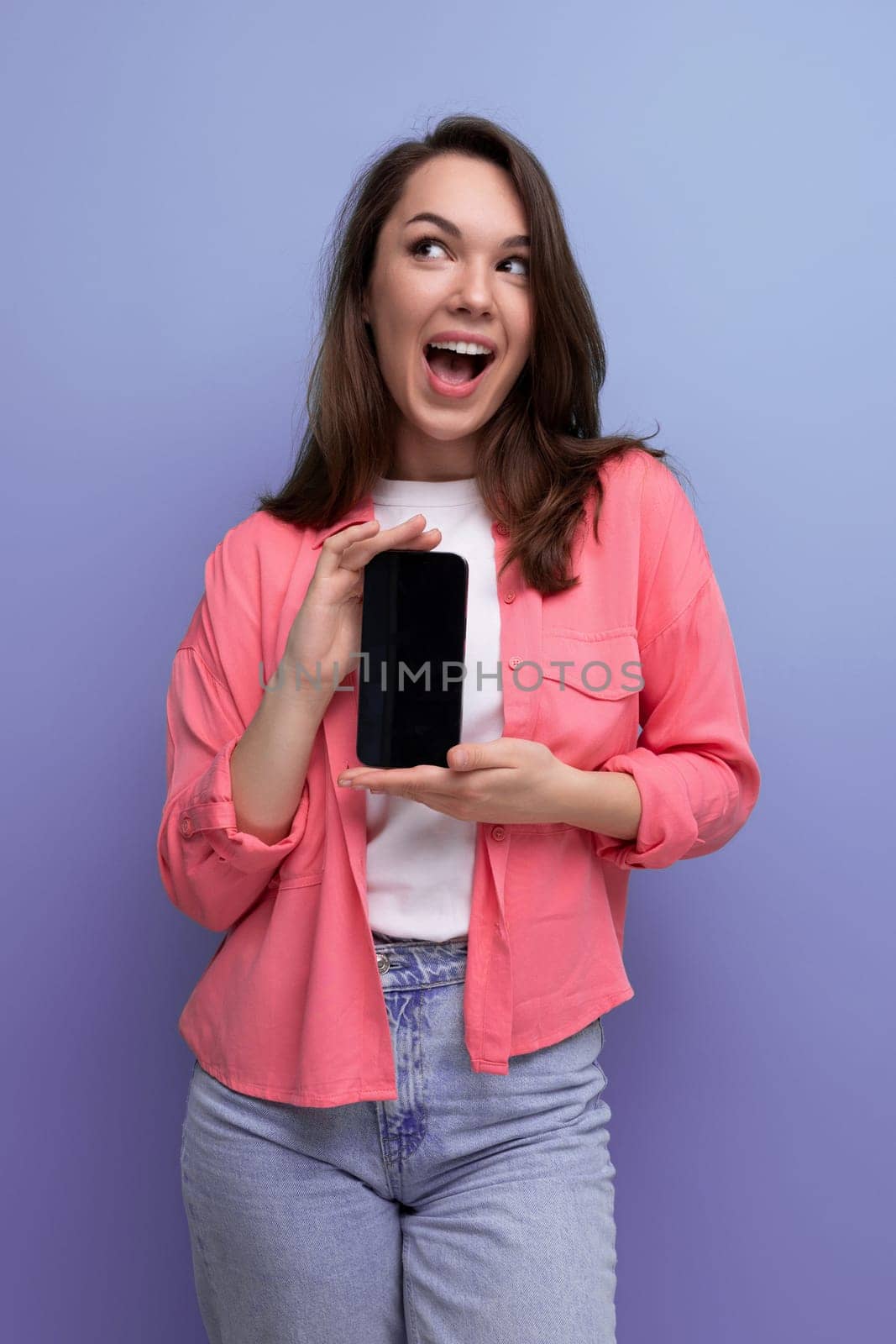 a cute brunette lady with dark hair below her shoulders in a shirt and jeans demonstrates a smartphone screen forward.