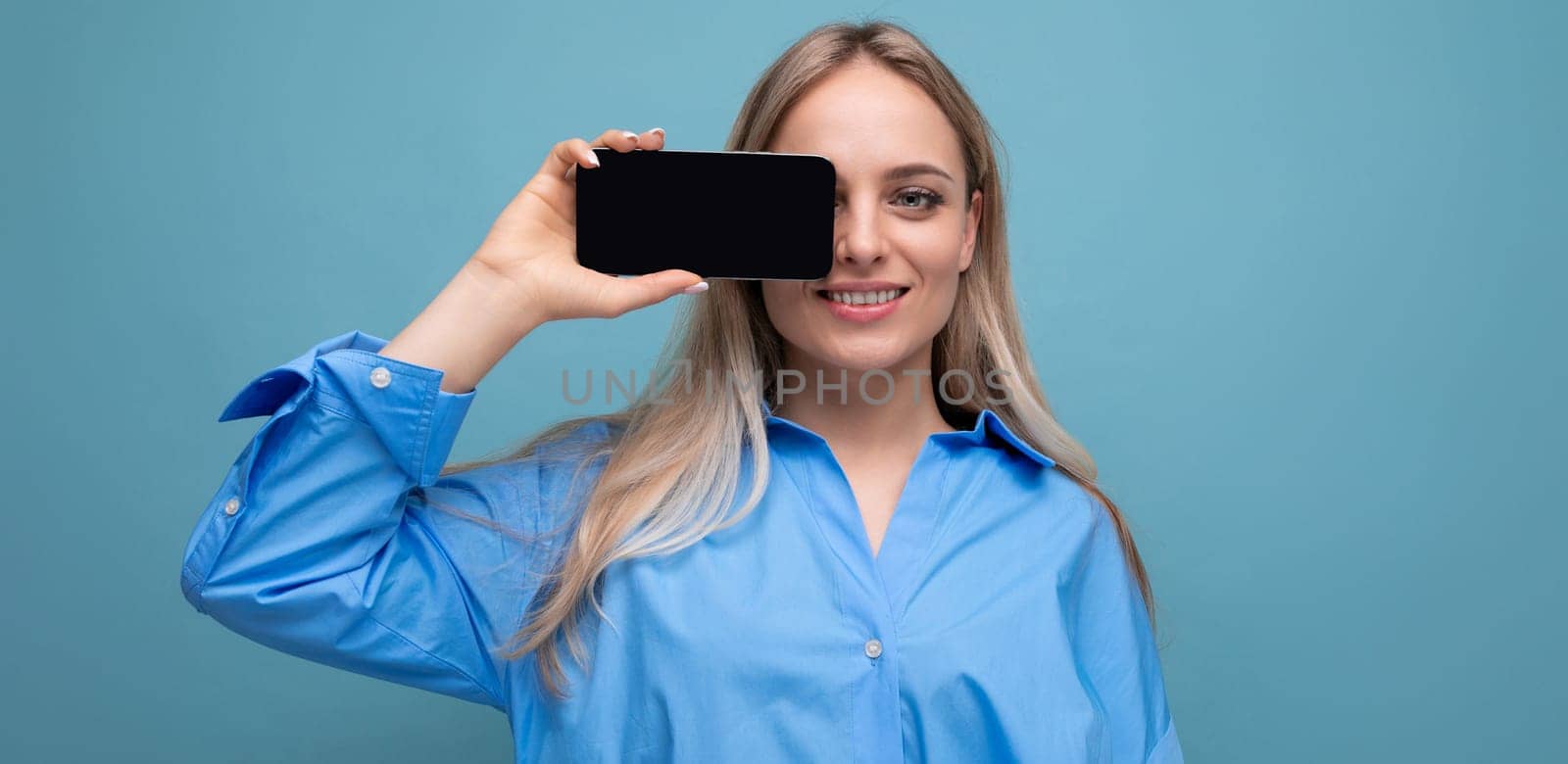 attractive confident blond girl holding a smartphone with a screen at the camera with a blank mockup for a website on a blue background by TRMK