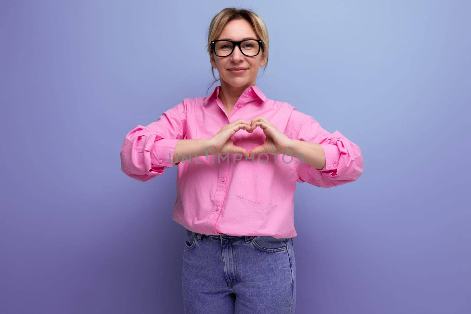 young energetic caucasian blonde secretary woman with ponytail hairstyle, glasses and in a pink shirt shows a heart on a studio background with copy space.