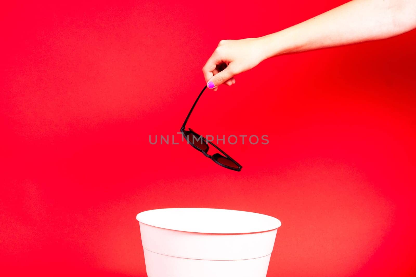 Hand throws sunglasses into trash bin with package on red background by Zelenin