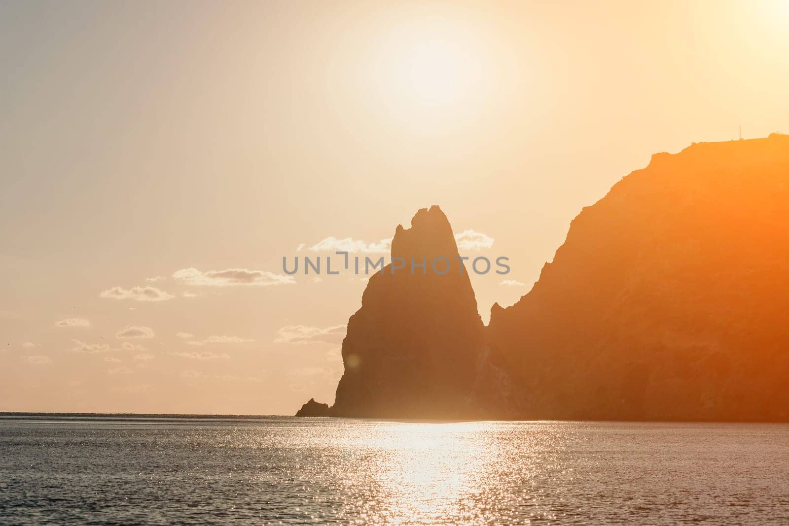 A red burning sunset with the silhouette of a cliff over the sea. Abstract nature summer or spring ocean sea background. Small waves on golden warm water surface with bokeh lights from sun.