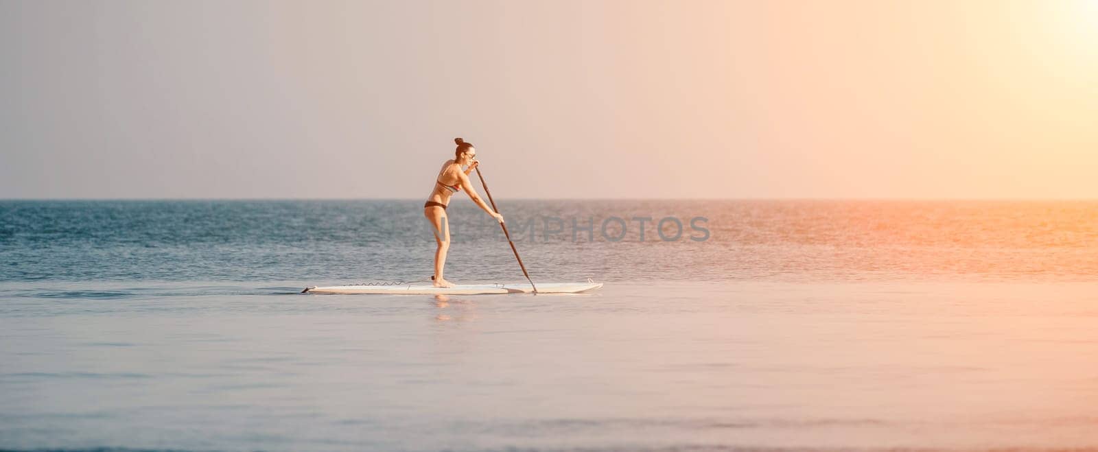 Sea woman sup. Silhouette of happy middle aged woman in rainbow bikini, surfing on SUP board, confident paddling through water surface. Idyllic sunset. Active lifestyle at sea or river. by panophotograph