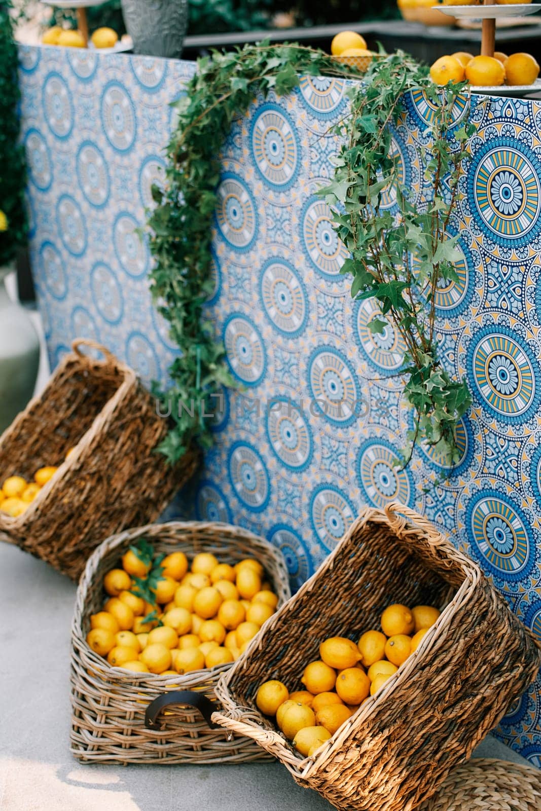 Wicker baskets with tangerines and lemons stand near a table covered with a colorful tablecloth and entwined with ivy. High quality photo