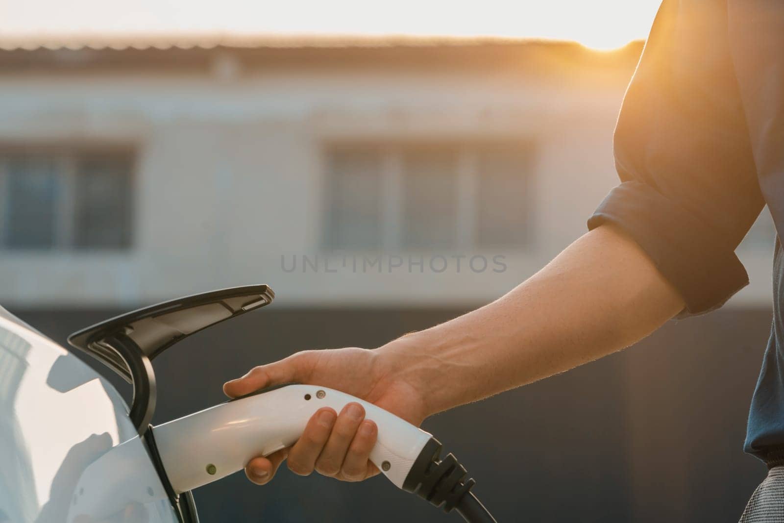 Young man put EV charger to recharge electric car's battery from charging station in city commercial parking lot with sunlight. Rechargeable EV car for sustainable eco friendly urban travel. Expedient