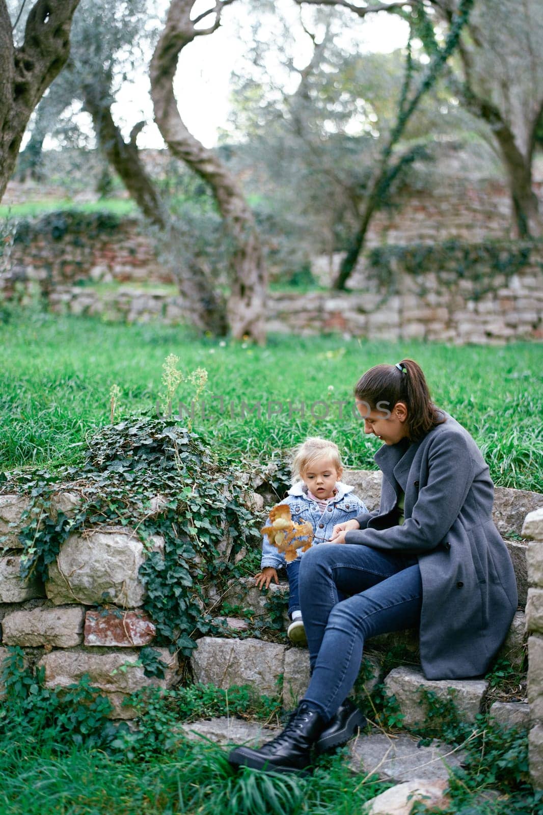 Mom with a little girl sits on stone steps in the garden and shows her a yellow branch. High quality photo
