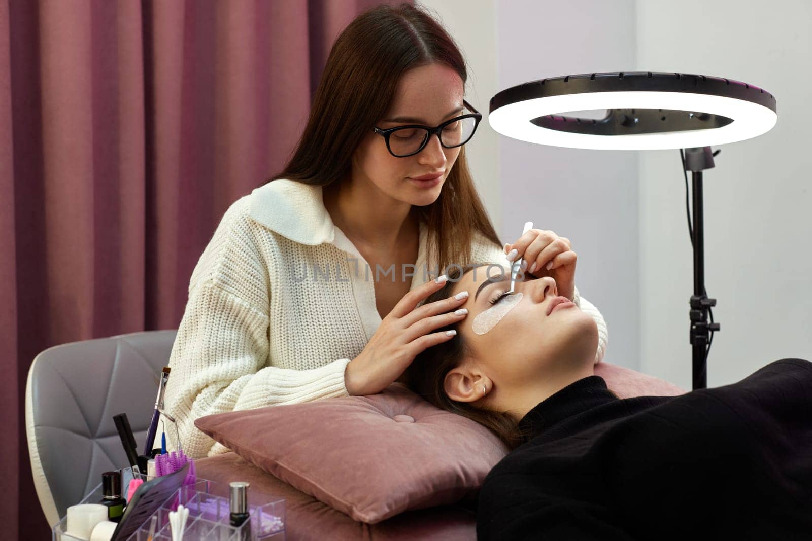 beautician with tweezers makes eyelash extension for young woman in beauty salon.