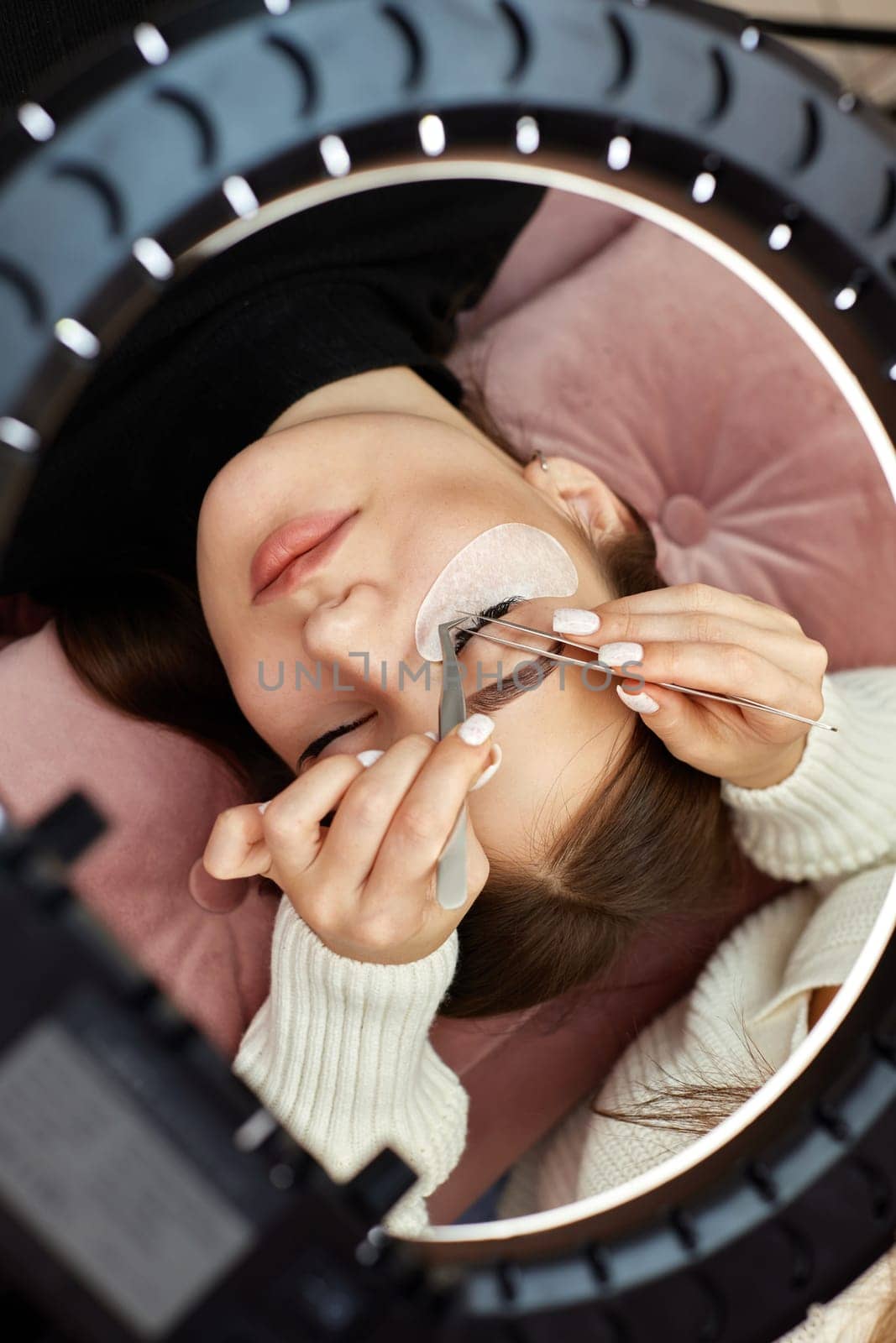 professional beautician with tweezers makes eyelash extension for beautiful woman under ring light lamp, top view