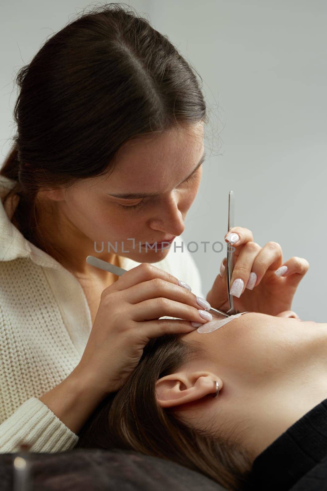 professional beautician with tweezers makes eyelash extension for beautiful woman in beauty salon.