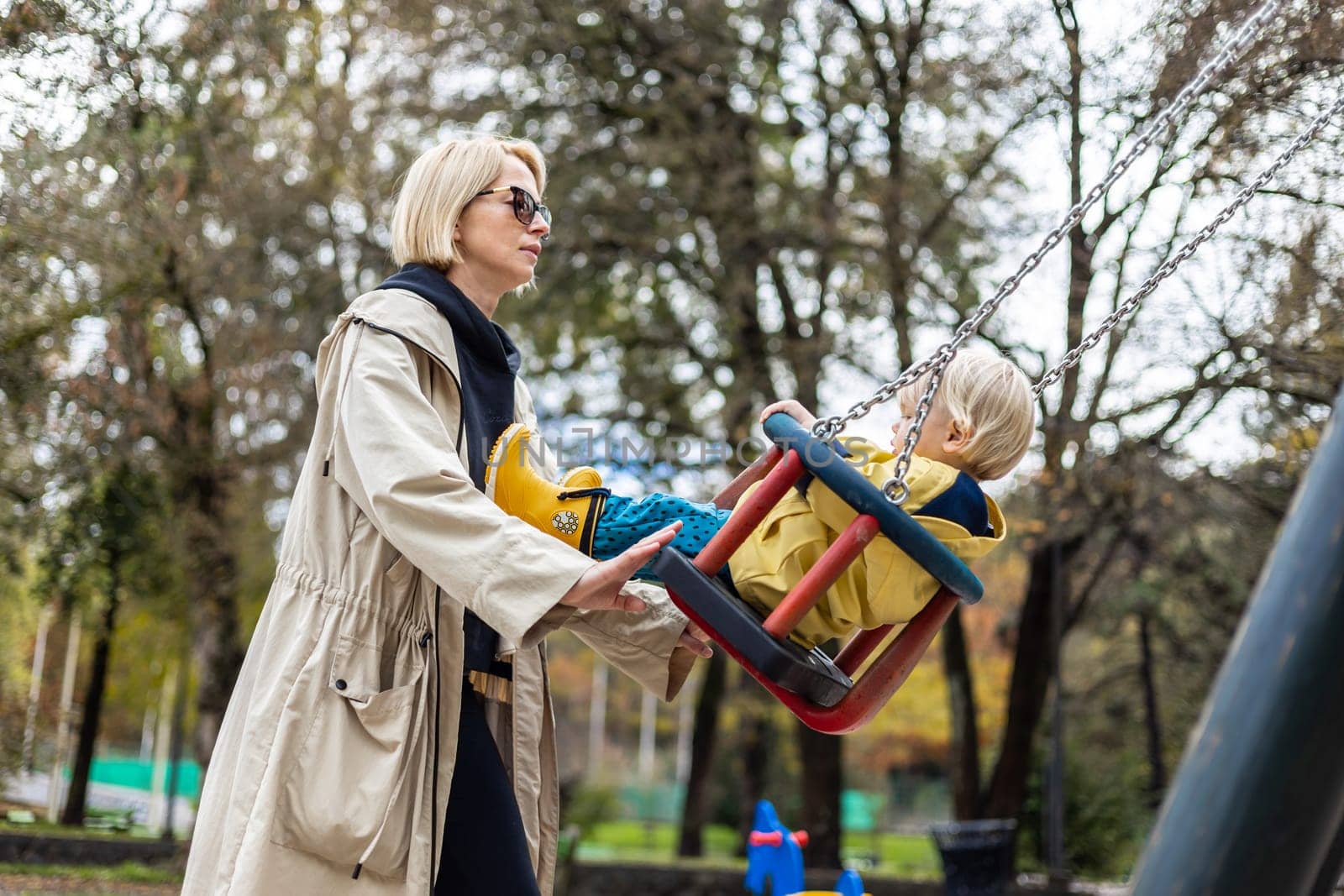 Mother pushing her infant baby boy child wearing yellow rain boots and cape on swing on playground outdoors on cold rainy overcast autumn day in Ljubljana, Slovenia by kasto