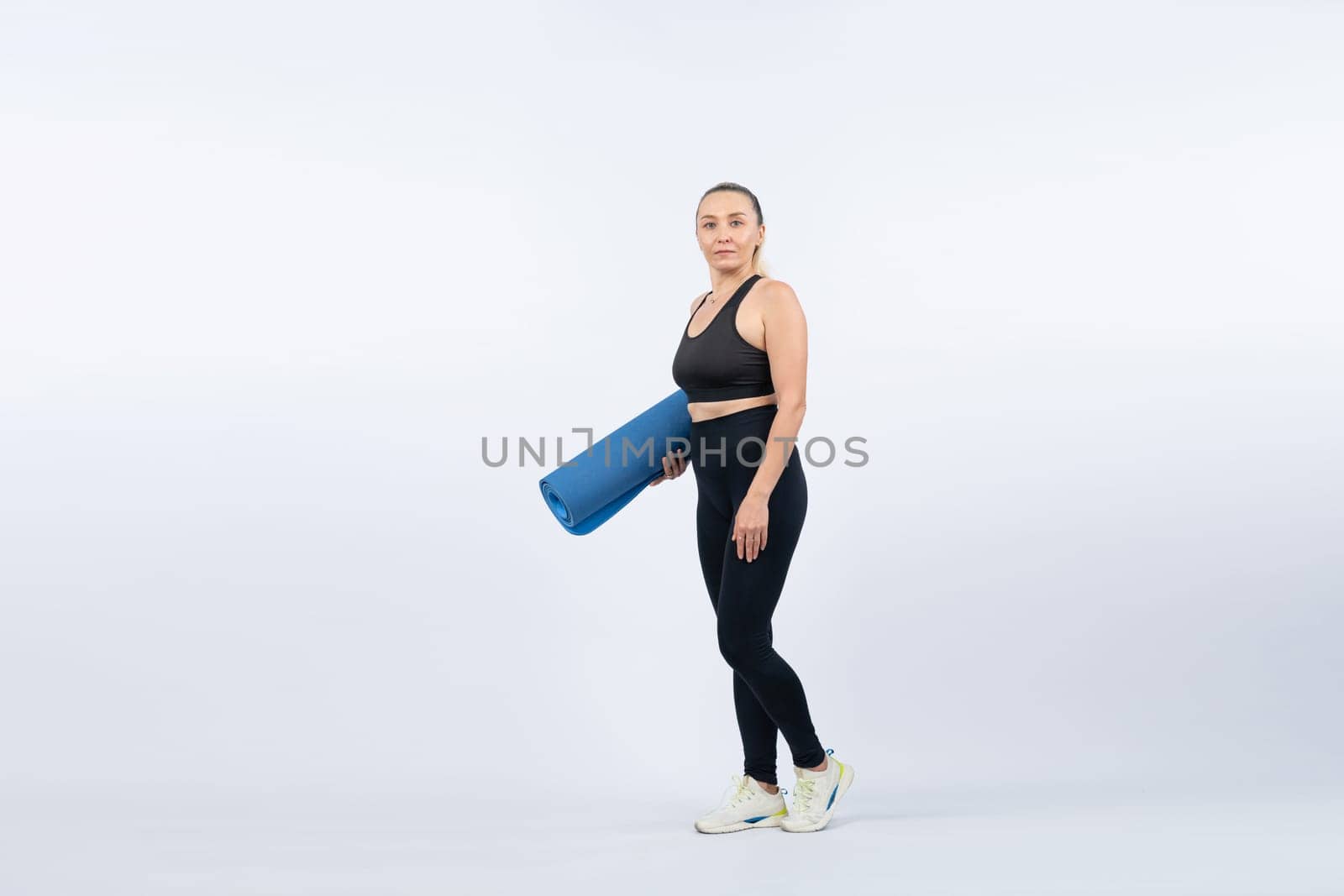 Full body length shot athletic and sporty senior woman holding fitness exercising mat on isolated background. Healthy active physique and body care lifestyle after retirement. Clout