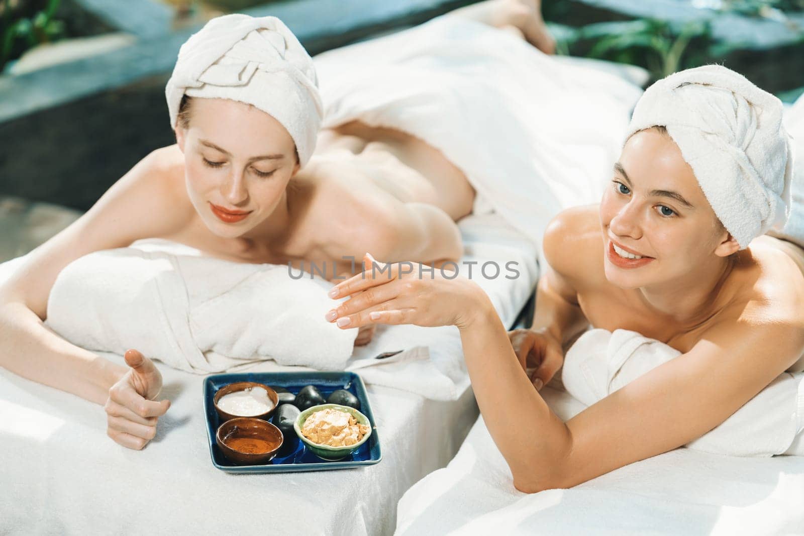 Couple of beautiful young girls lie on spa bed during interested in homemade beauty facial mask. Attractive woman touching herbal facial mask. Surrounded with nature environment. Tranquility.