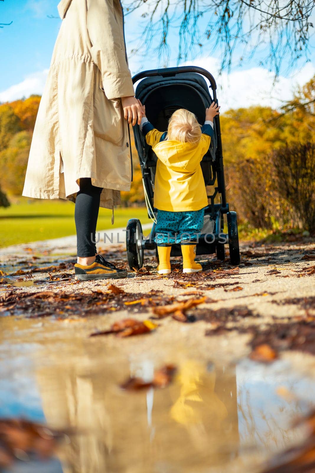 Sun always shines after the rain. Small blond infant boy wearing yellow rubber boots and yellow waterproof raincoat walking in puddles, pushing stroller in city park, holding mother's hand after rain