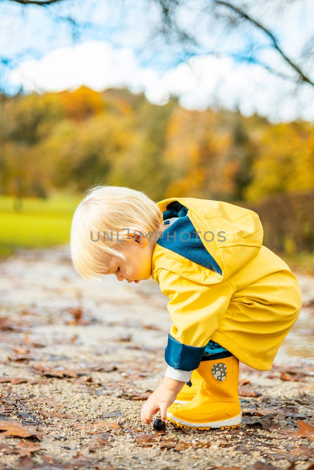 Sun always shines after the rain. Small bond infant boy wearing yellow rubber boots and yellow waterproof raincoat walking in puddles in city park on sunny rainy day. by kasto