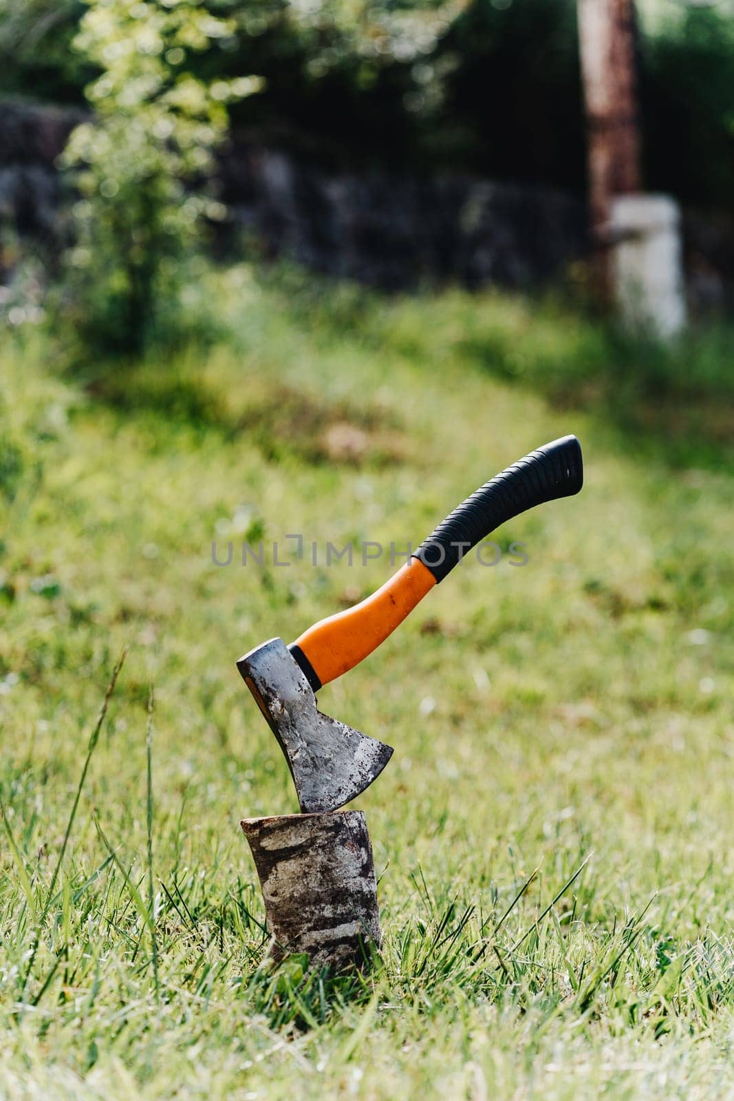 Axe with orange handle sticks out in hemp against the background of green grass by Praximon