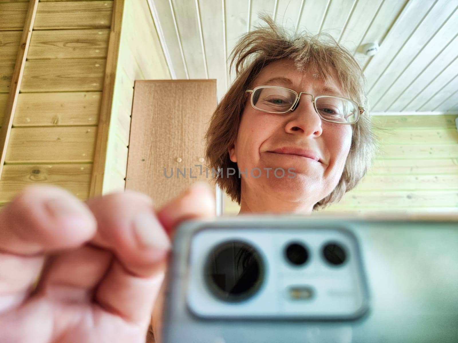 A shaggy, funny middle-aged woman in glasses takes selfie on her smartphone in front of the mirror in the morning. A blogger girl with disheveled hair takes pictures of herself for a blog