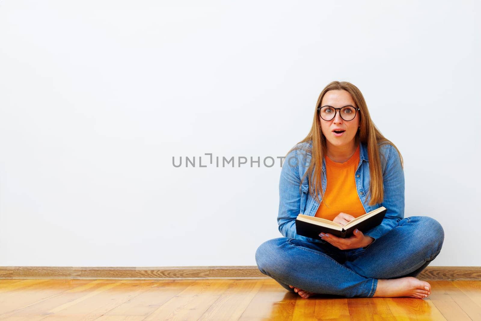 Surprised woman with glasses sits on the floor against white wall holding book by andreyz