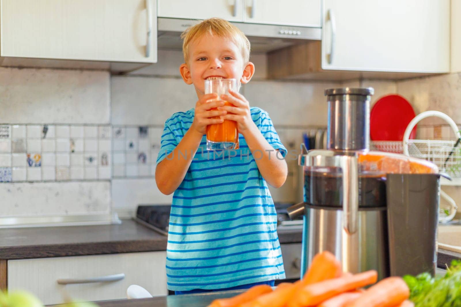 Little boy drinking freshly squeezed carrot juice from a glass in the kitchen. Concept of healthy and delicious food