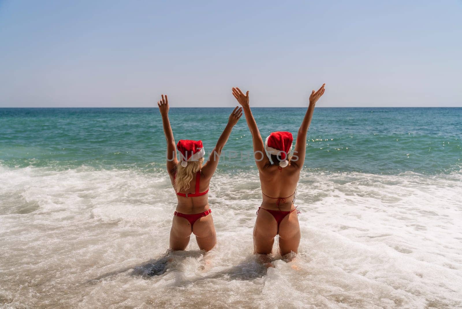 Women Santa hats ocean play. Seaside, beach daytime, enjoying beach fun. Two women in red swimsuits and Santa hats are enjoying themselves in the ocean, kneeling in the waves and raising their hands up. by Matiunina