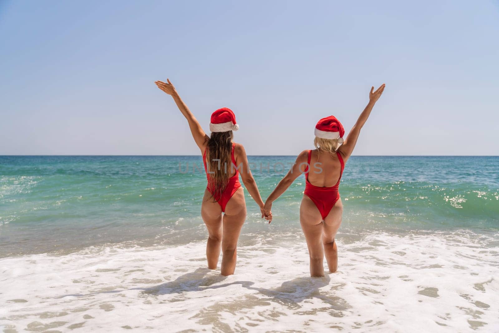 Women Santa hats ocean play. Seaside, beach daytime, enjoying beach fun. Two women in red swimsuits and Santa hats are enjoying themselves in the ocean waves and raising their hands up