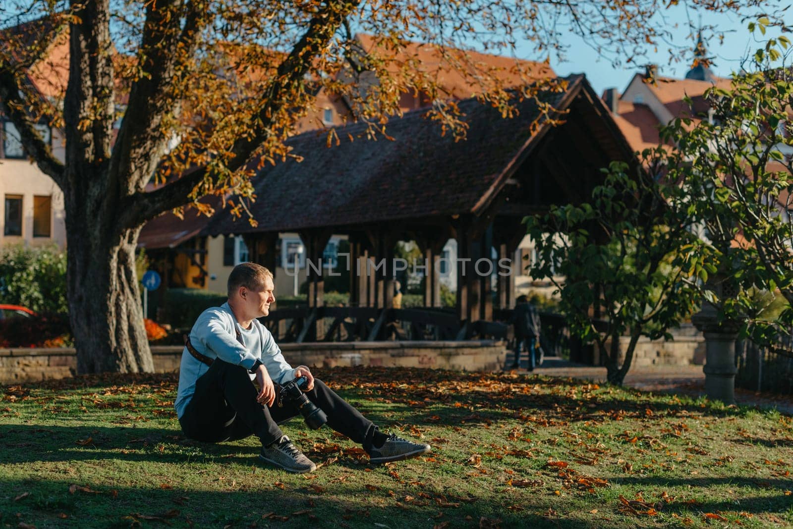 Professional Photographer Taking Picture Of Beautiful Autumn Park. Man Professional Photographer Sit With Camera And With Smartphone In Autumn Park. Retouched, Vibrant Colors, Brownish Tones. by Andrii_Ko