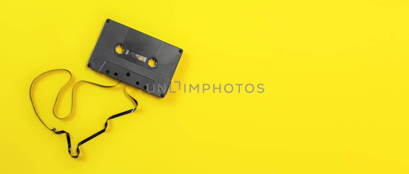 Overhead shot - magnetic audio cassette with some tape out, on yellow board, wide banner space for text on right side. by Ivanko