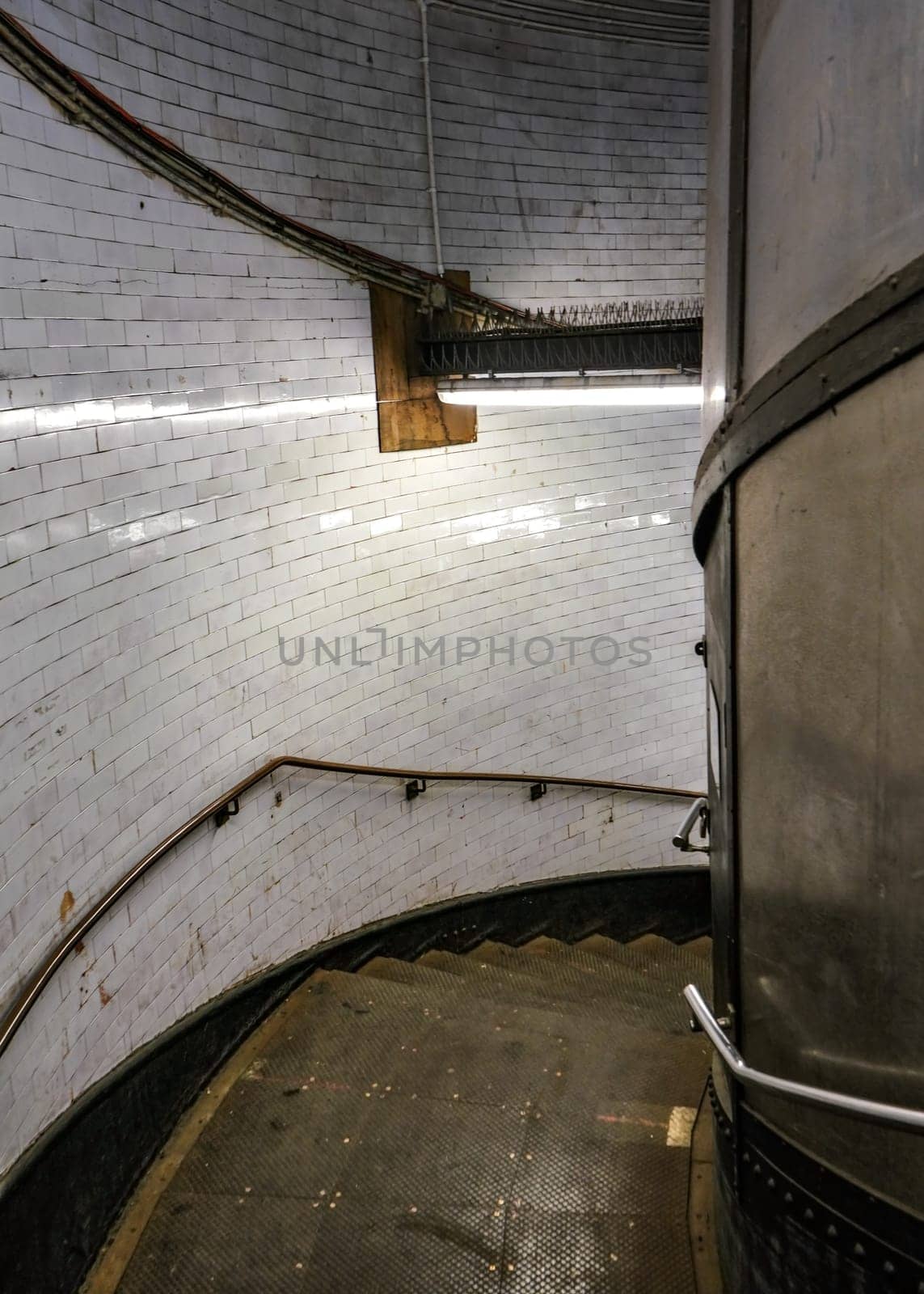 Dark and dirty metal spiral staircase lit by neon light, leading down to Greenwich Foot Tunnel under river Thames in London by Ivanko