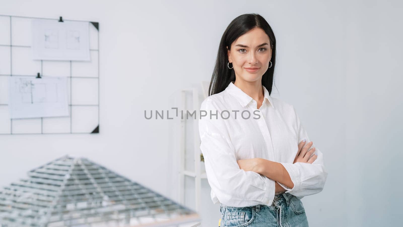 Close up portrait of young beautiful professional architect engineer cross arms and poses confidently with house model at white modern office with blueprint. Creative living and design. Immaculate.