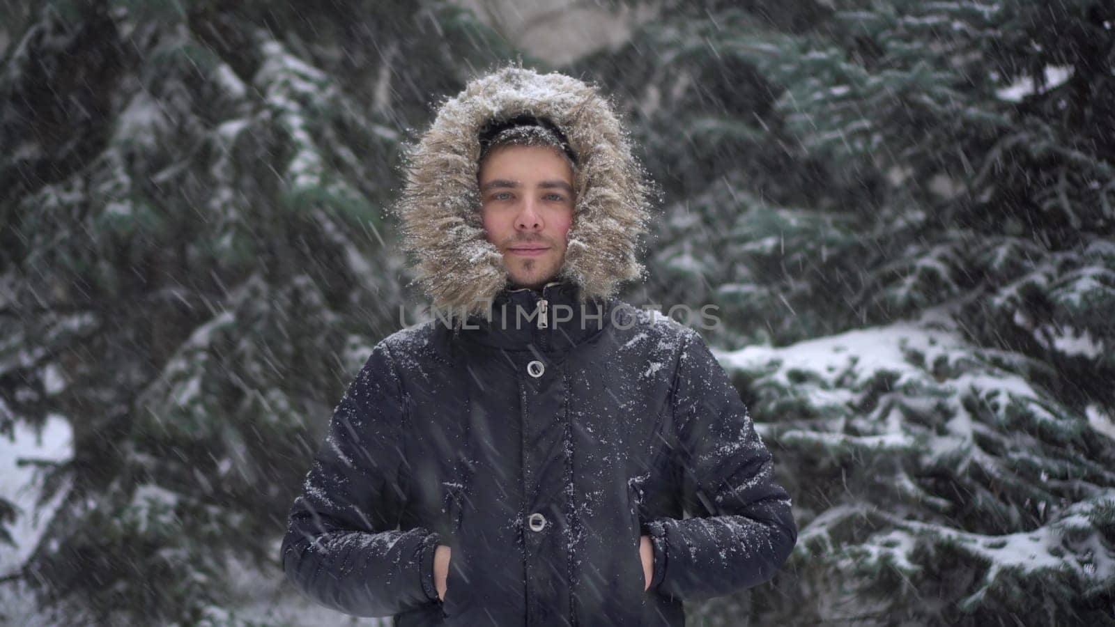 A young man stands against a background of fir trees under heavy snowfall in winter. A man in a down jacket with a hood stands and looks at the camera. by Puzankov