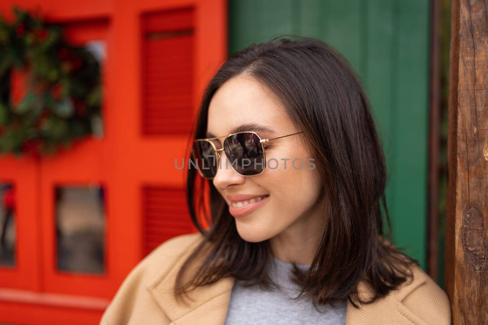 Smiling woman wear sunglasses close up portrait on Christmas market. Woman dressed in coat with sunglasses leaning on pole outside Christmas store. Female standing on street