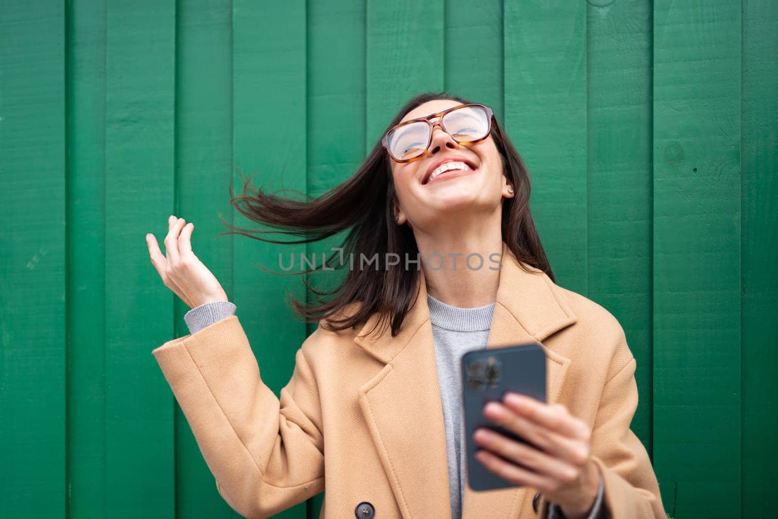 Woman with smartphone leaning against wall fixes hair hold phone in hand. Portrait of happy smiling adult young woman dressed stylish beige coat standing outdoor. Positive emotions