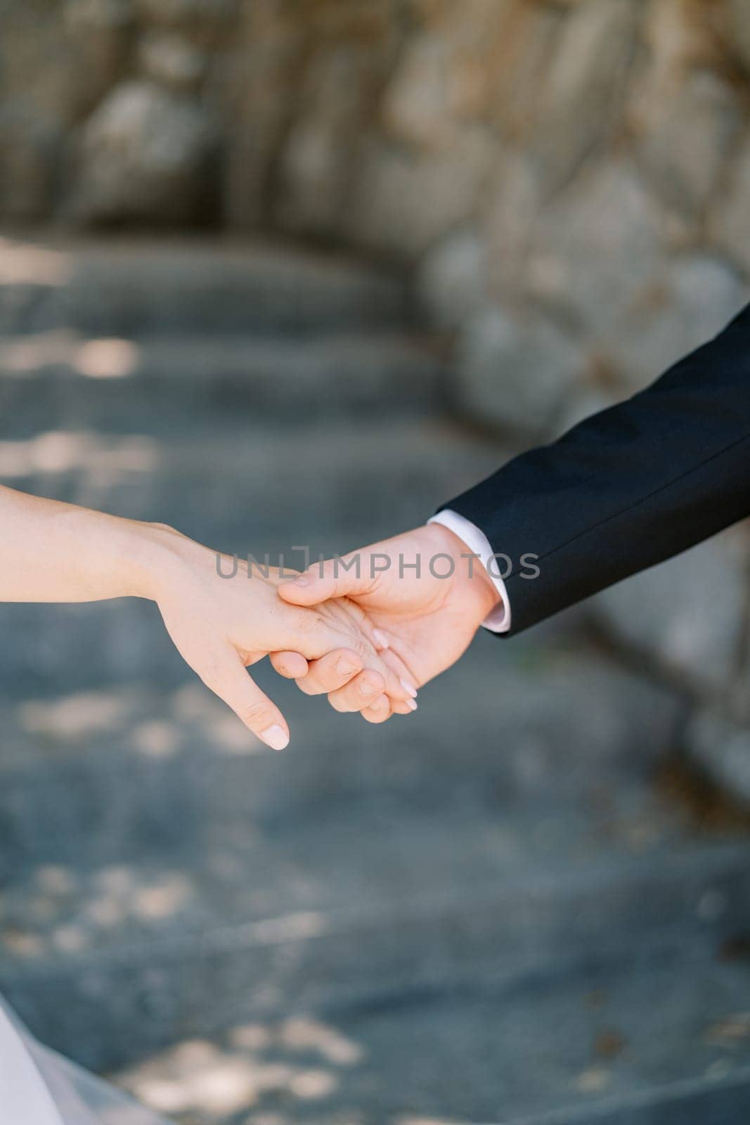 Groom holds bride hand while standing at the stone steps in the garden. Cropped. Faceless by Nadtochiy