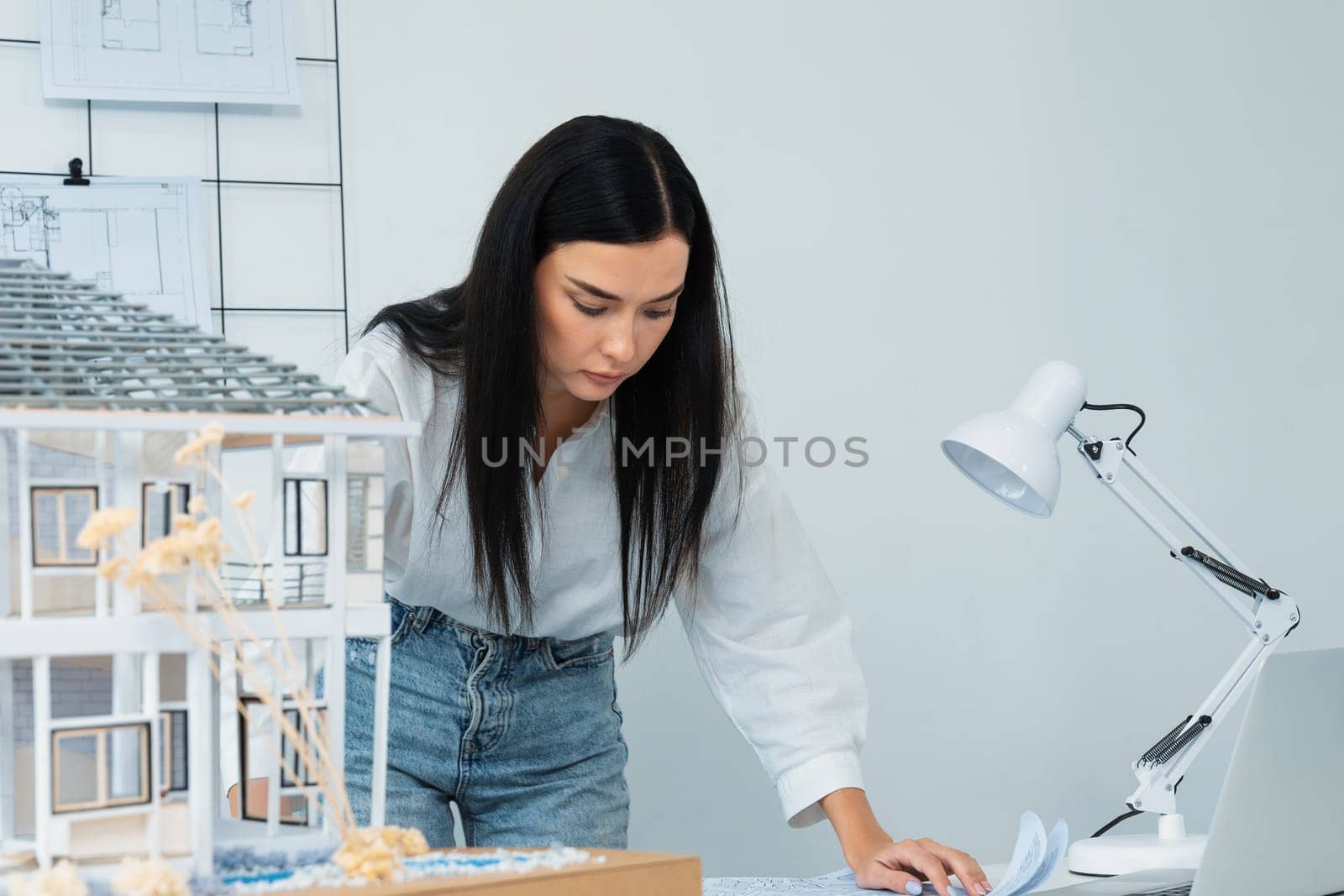 Portrait of Young beautiful caucasian architect focus on comparing house structure between house model and blueprint and laptop placed on meeting table. Check, inspect, estimate concept. Immaculate.