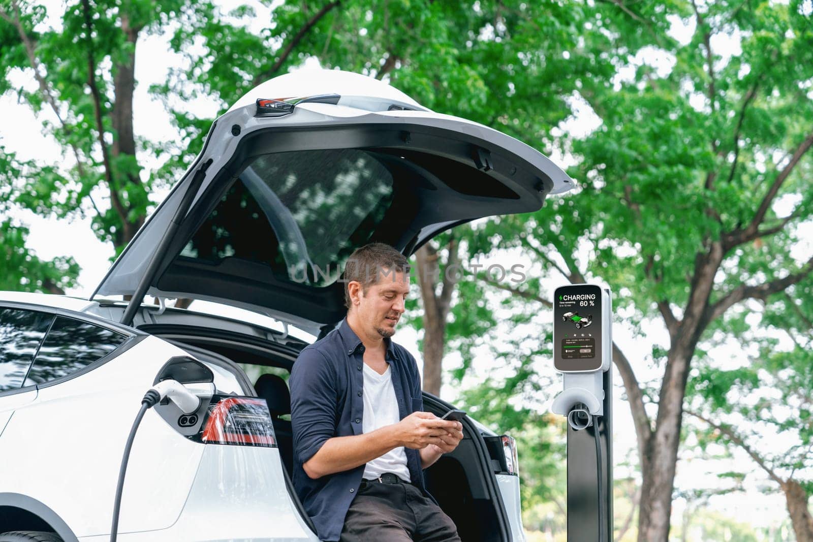 Man using smartphone online banking application to pay for electric car battery charging from EV charging station during vacation holiday road trip at national park or summer forest. Exalt