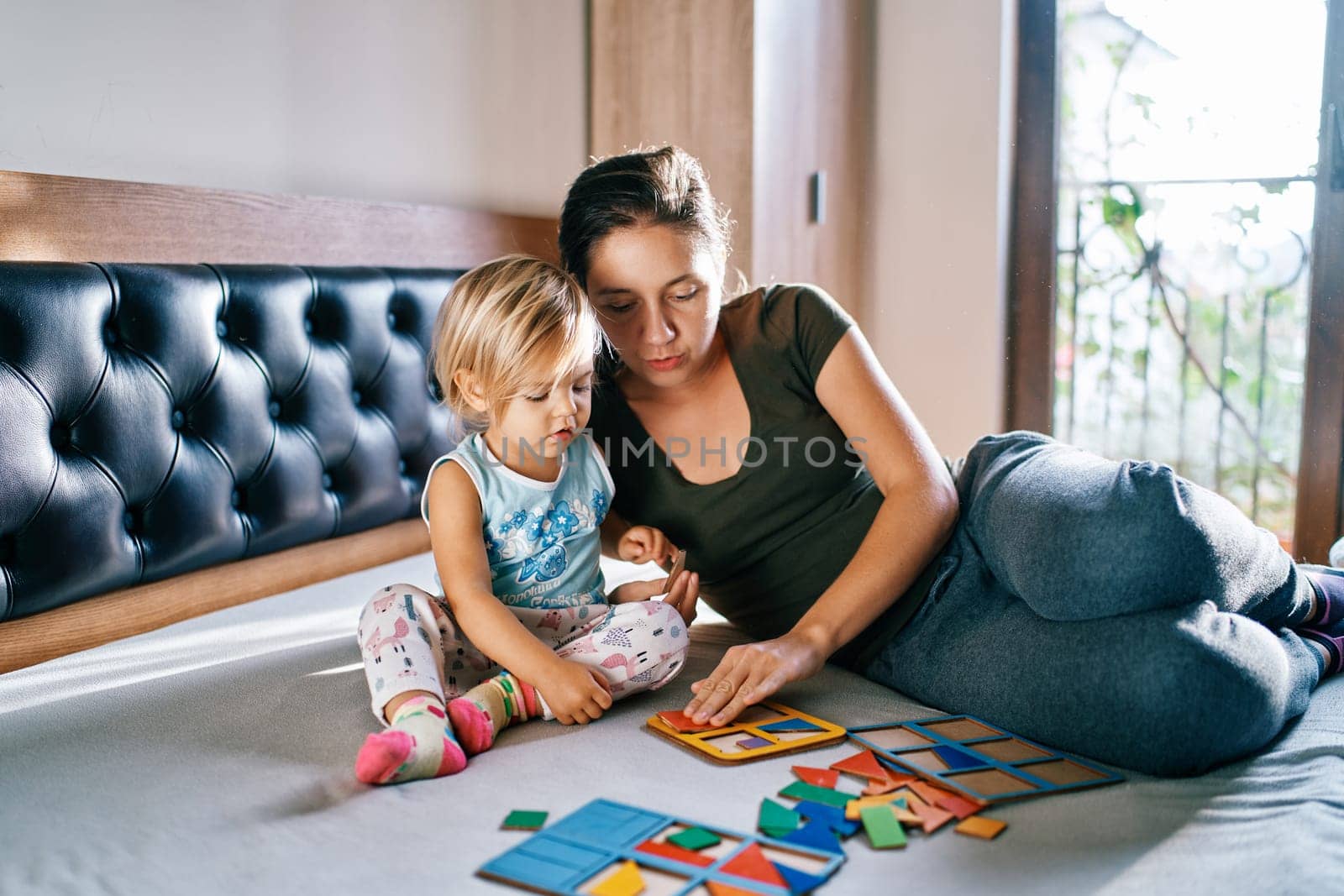 Mom teaches a little girl to collect puzzles in a cardboard tablet while sitting on the bed by Nadtochiy