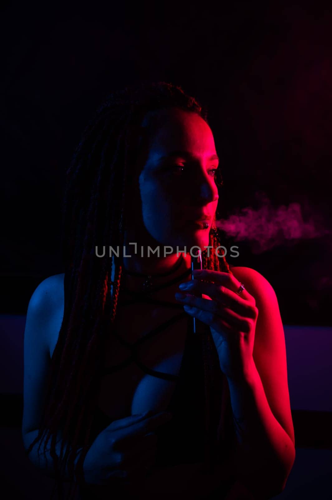 Profile of caucasian girl with dreadlocks smokes a vape in red blue light