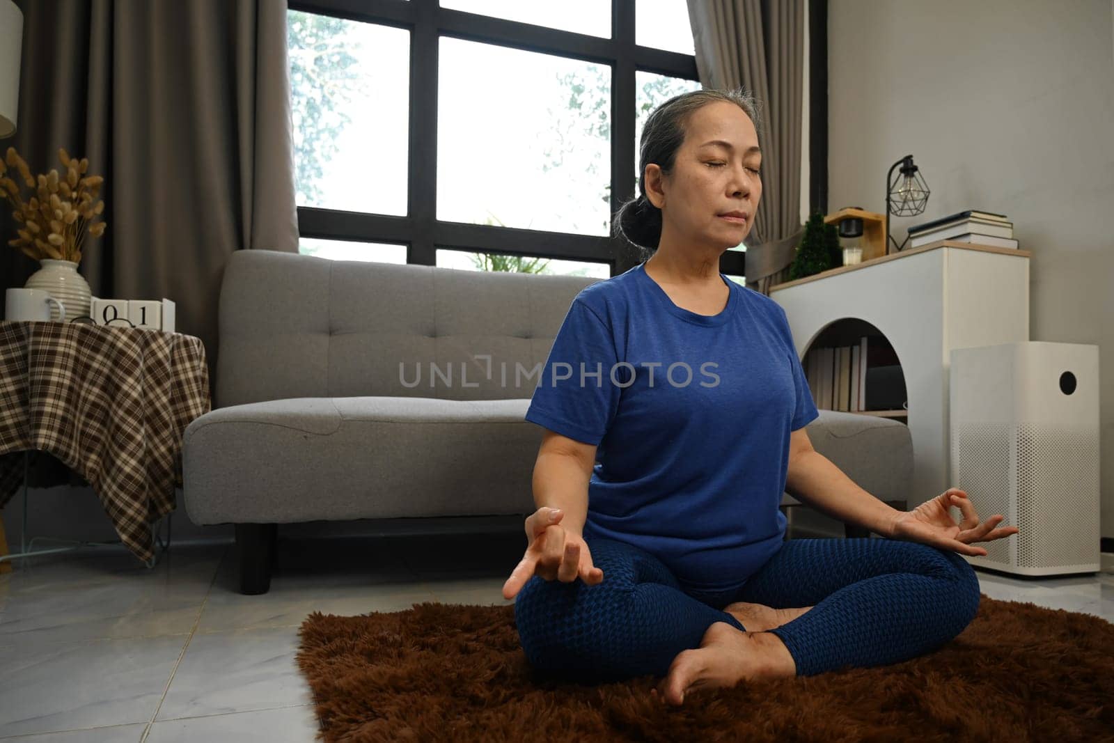 Serene senior woman meditating at home sitting in lotus pose. Active lifestyle and healthcare concept.