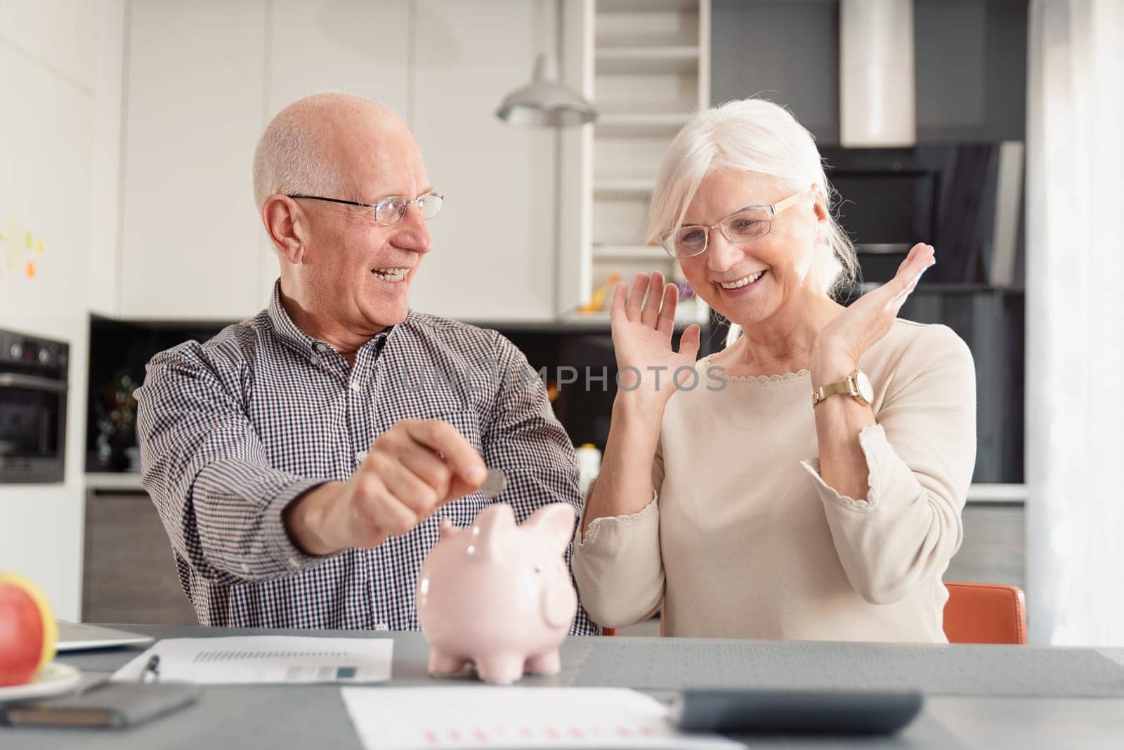Funny photo of mature couple putting coin into piggy bank at home. Savings, pension plan concept