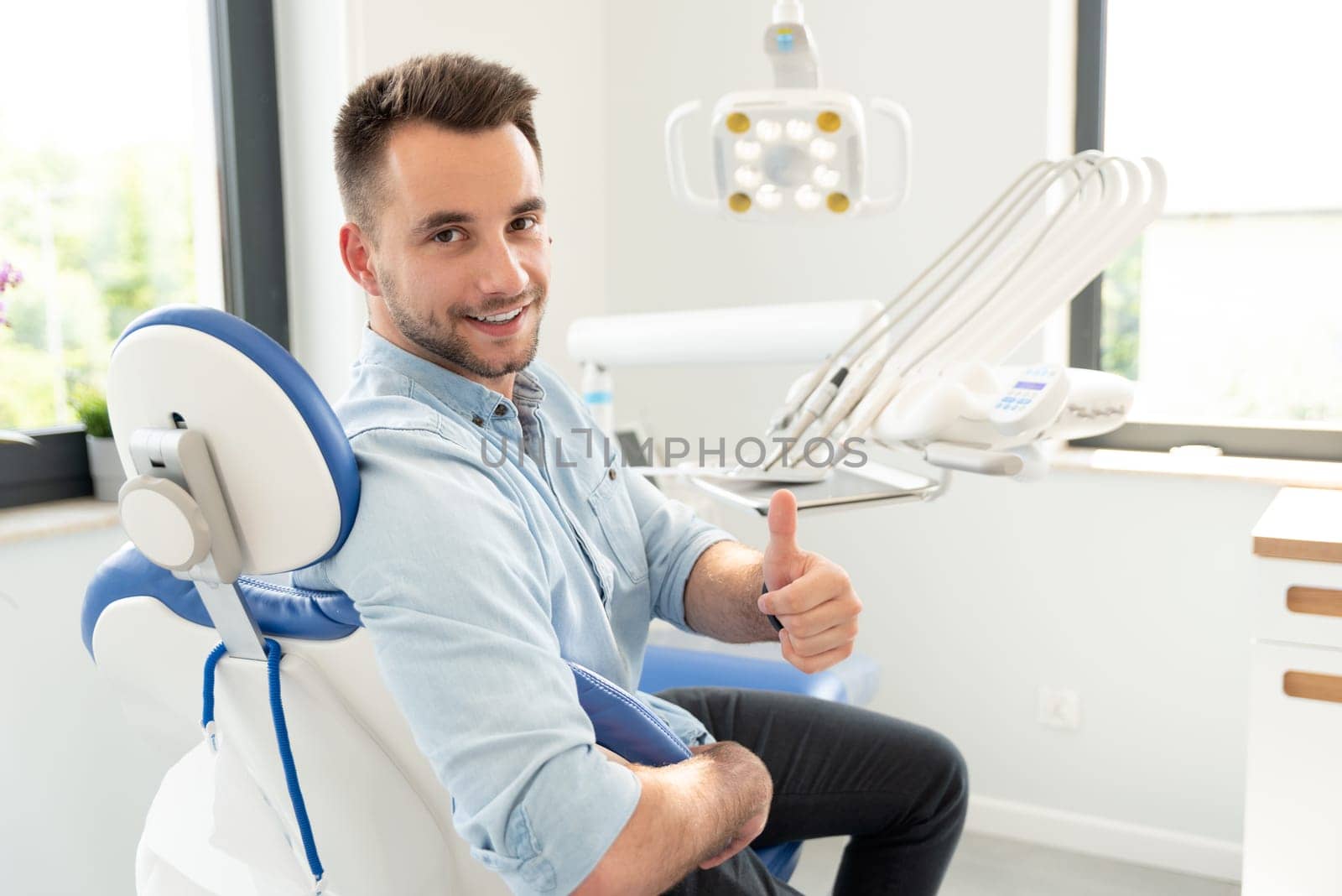 Handsome man showing thumbs up and smiling sitting at the dental chair. Professional dental clinic, healthy teeth concept.