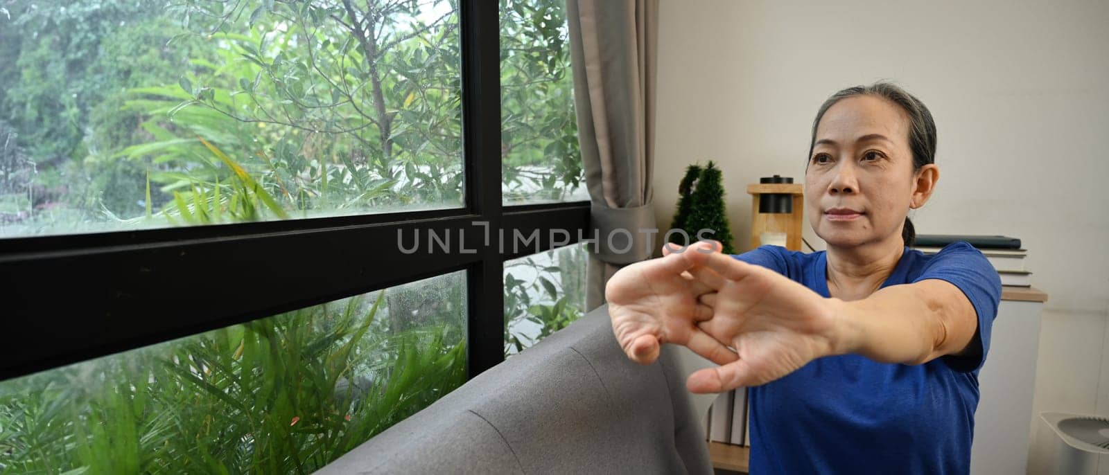 Beautiful senior lady stretching arms and looking away in living room. Sports and Healthy lifestyle concept.