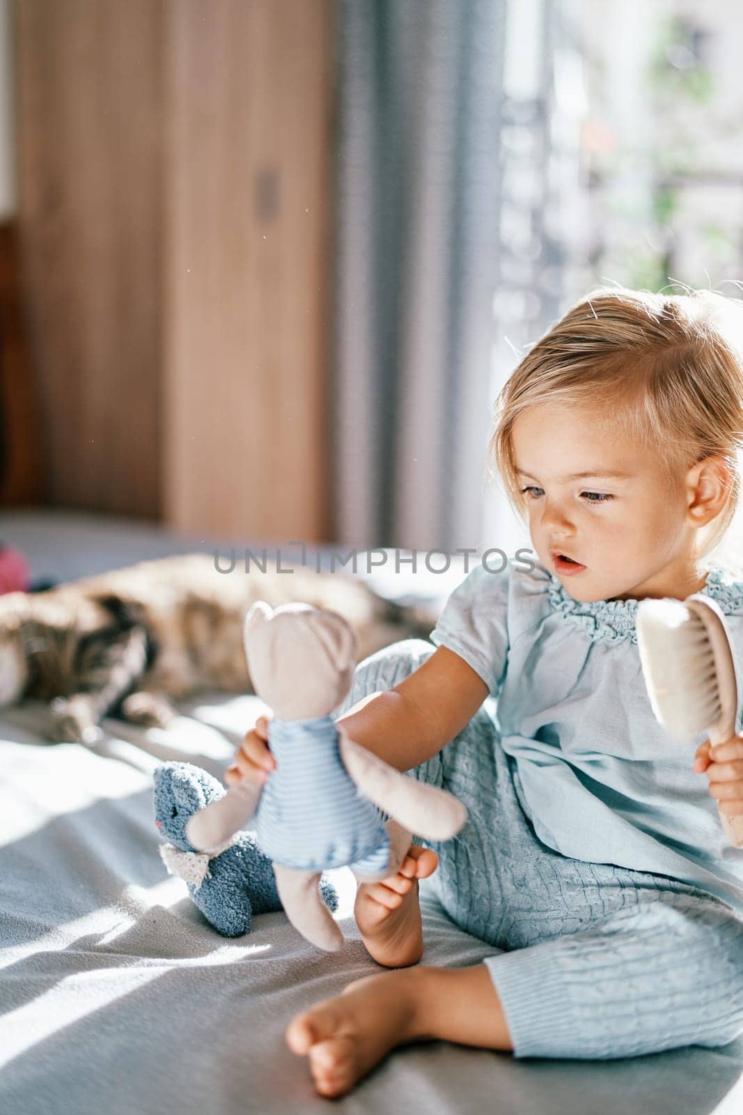 Little girl holds a teddy bear in her outstretched hand with a comb in her other hand while sitting on the bed. High quality photo