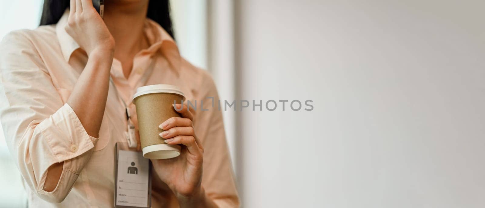 Young woman employee holding a paper cup of hot coffee and talking on mobile phone.