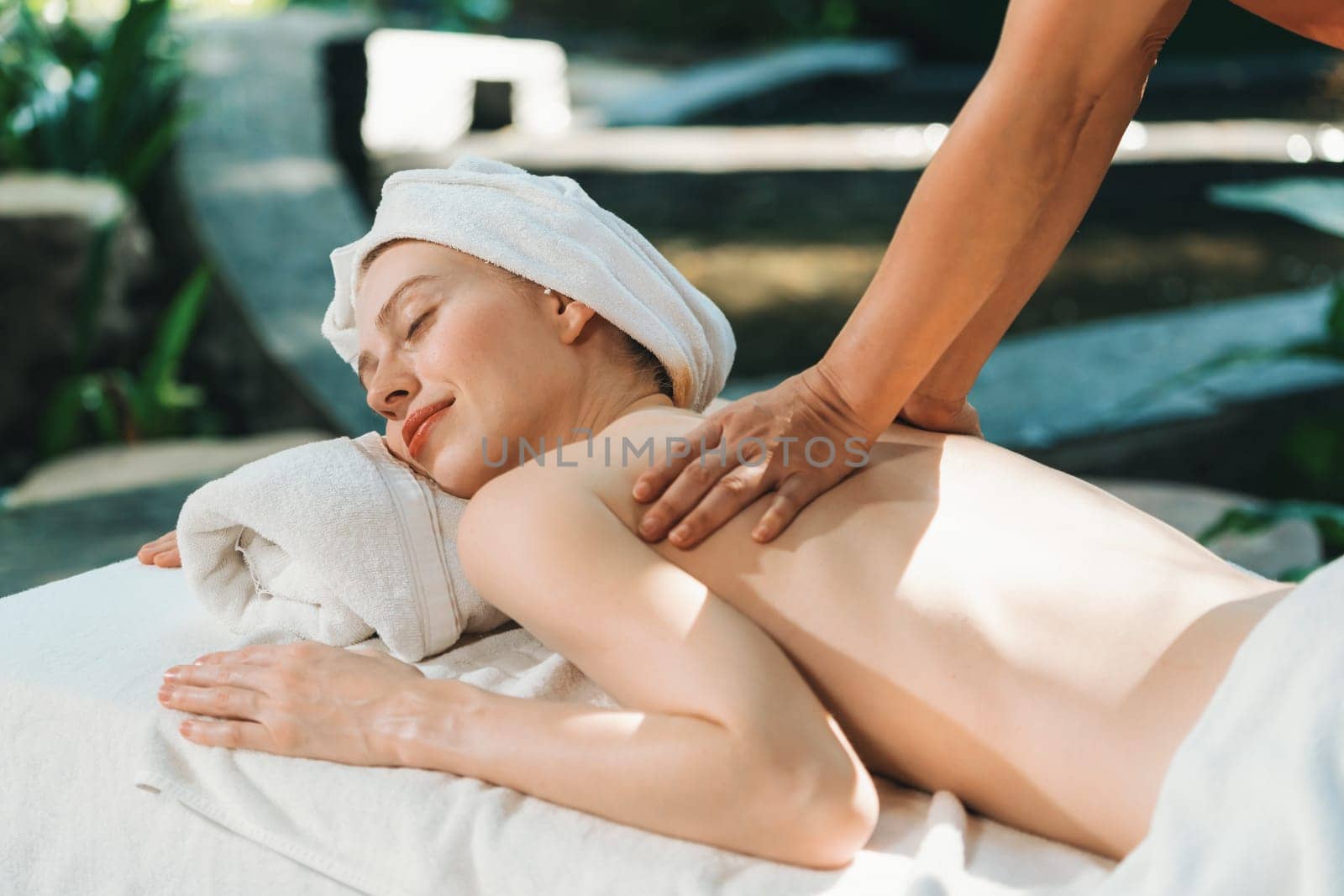 Beautiful young woman received a back massage on a spa bed from professional masseuse. Attractive female relaxes deeply by skilled hands of the massage therapist. Surrounded with nature. Tranquility.