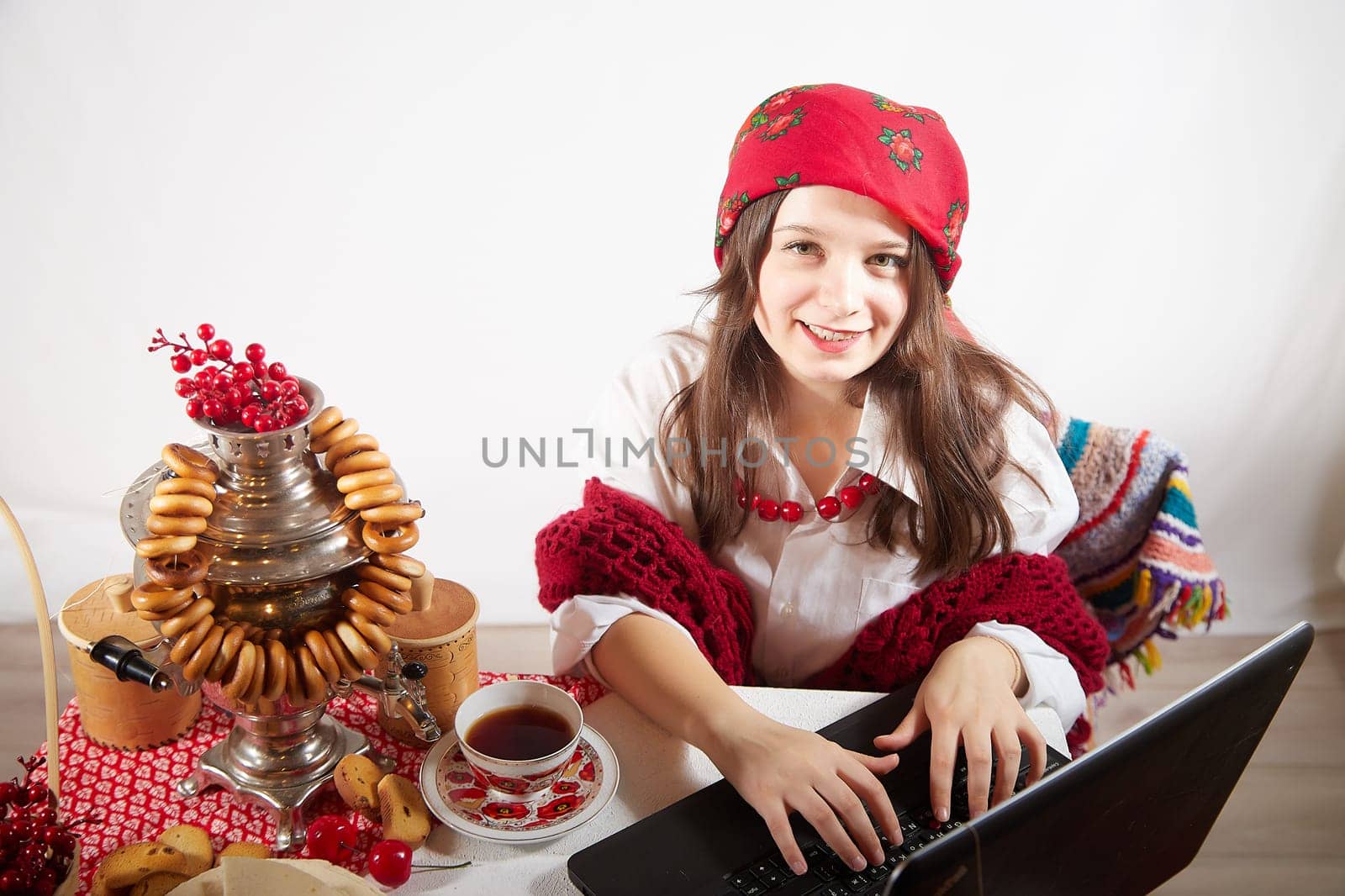 Girl in red colored scarf, white shirt and shawl near a table with a samovar, bagels and laptop in Easter holiday or Masolennitsa. A freelancer or blogger with a laptop is resting and working