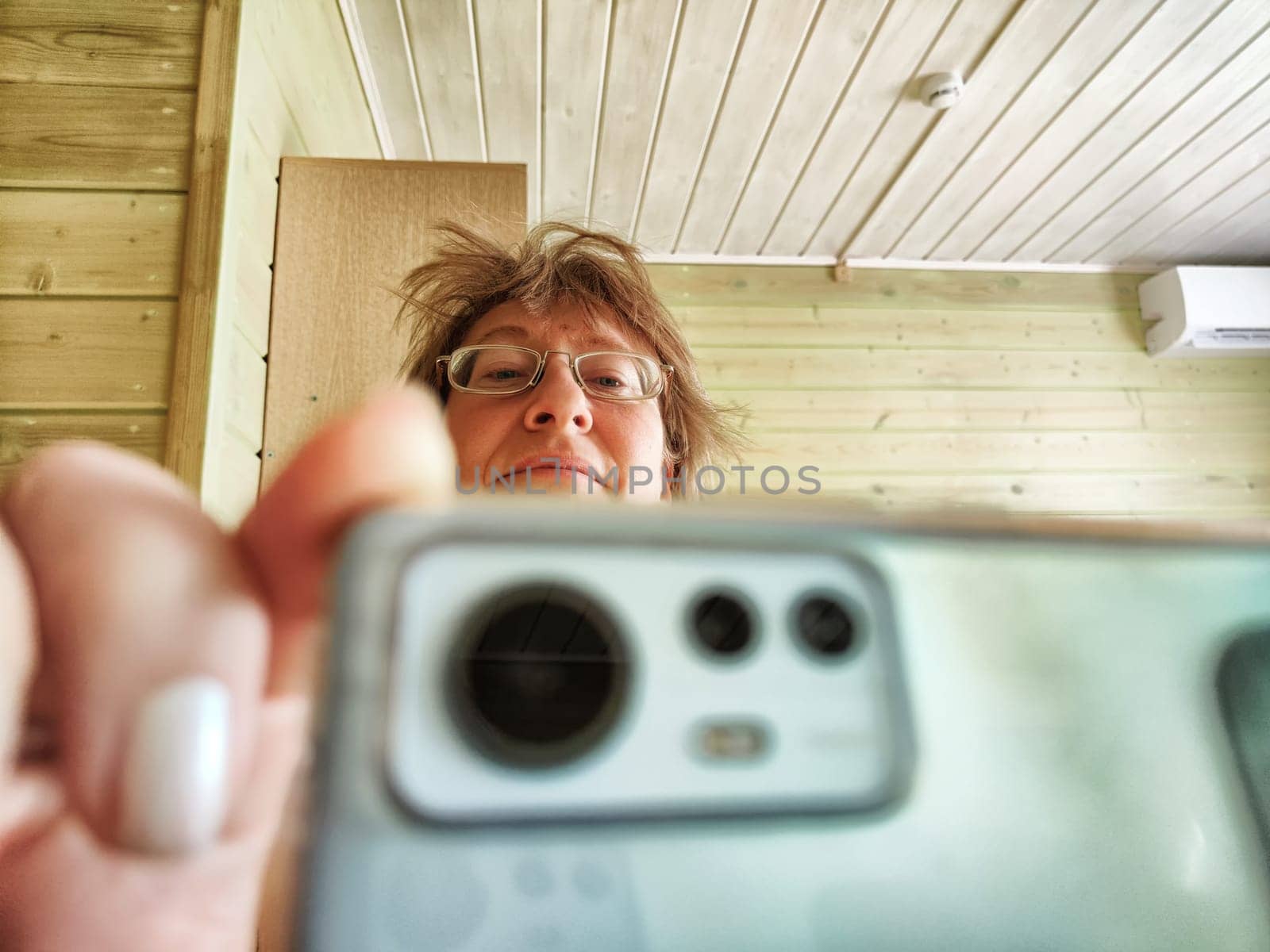 A shaggy, funny middle-aged woman in glasses takes selfie on her smartphone in front of the mirror in the morning. A blogger girl with disheveled hair takes pictures of herself for a blog