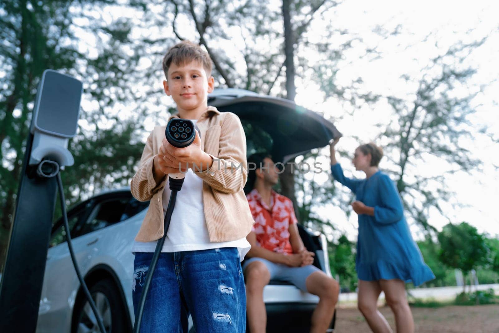 Little boy holding EV charger and point at camera with his family. Perpetual by biancoblue