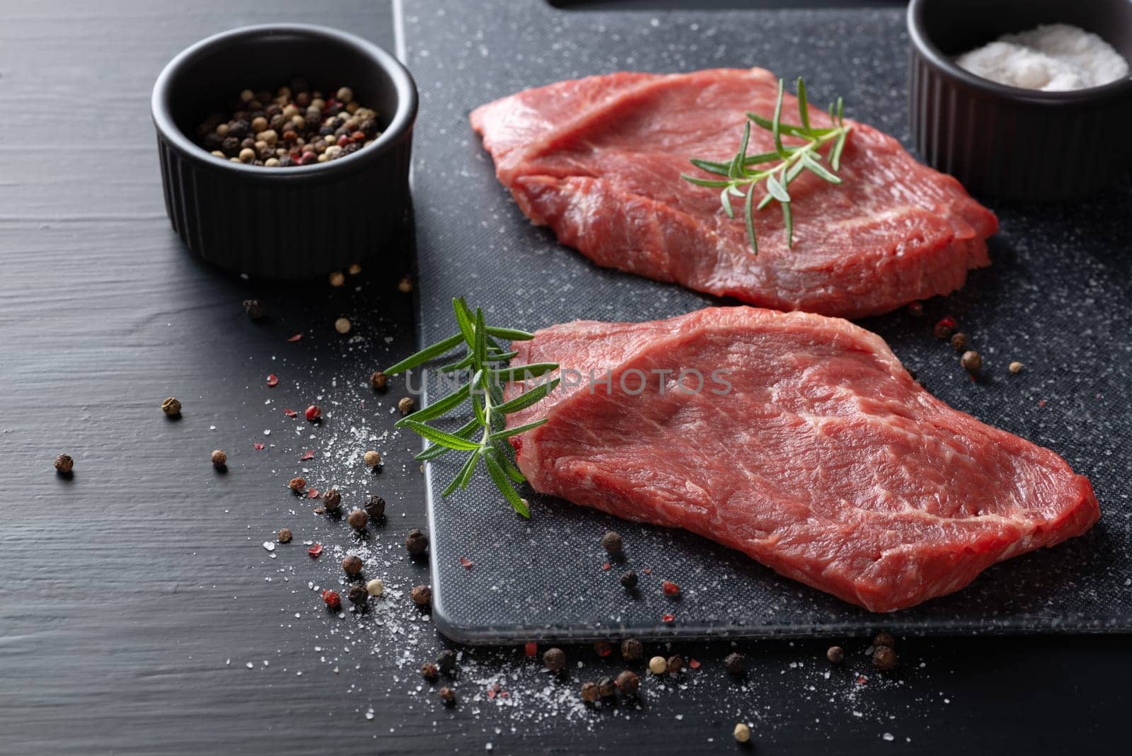 Two fresh raw marbled beef steaks on a cutting board on a wooden background with salt, pepper and rosemary in a rustic style.