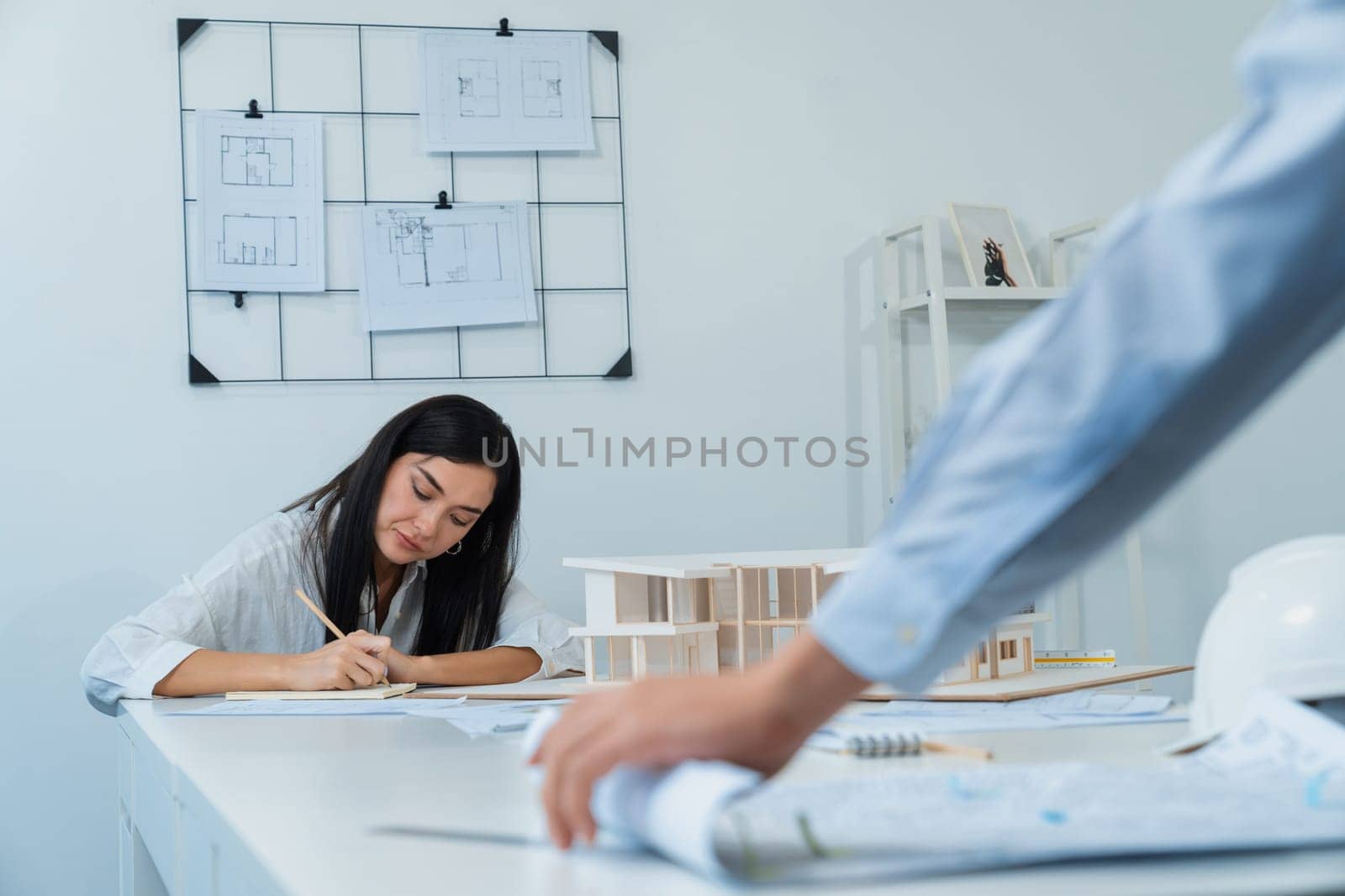 Professional asian male project manager hand puts the map on meeting table while beautiful colleague taking a note about house structure at meeting room with blueprint hanged behind. Immaculate.