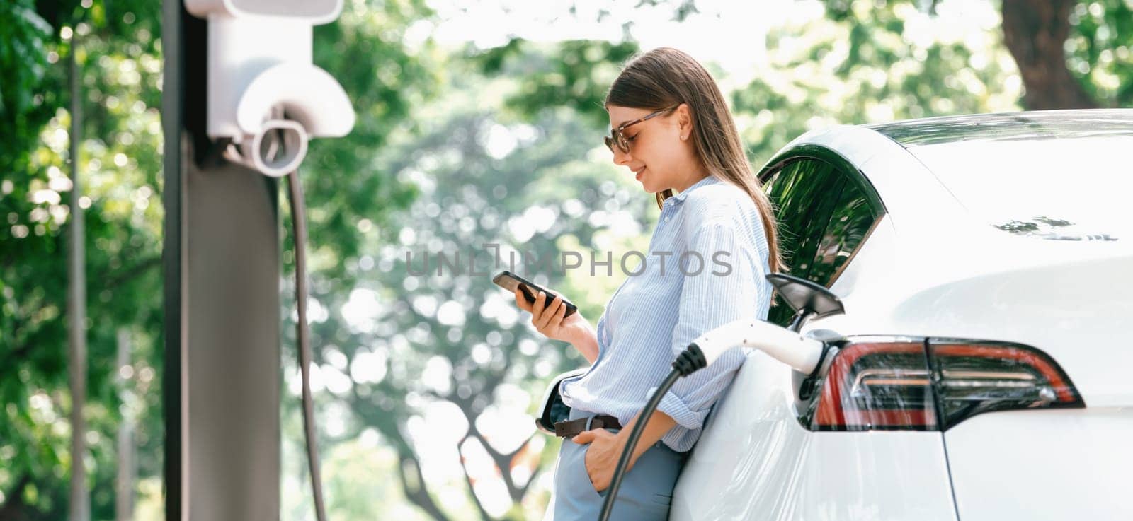 Young woman using smartphone online banking application to pay for electric car battery charging from EV charging station during vacation road trip at national park or summer forest. Panorama Exalt